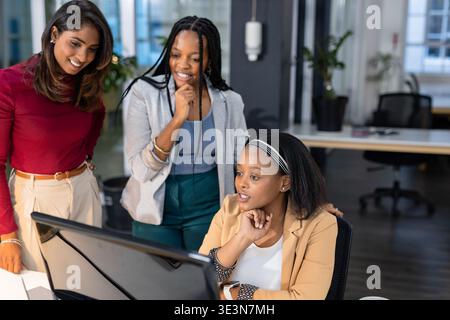 Diverse female colleagues collaborating around computer monitor in open office with potted plants Stock Photo