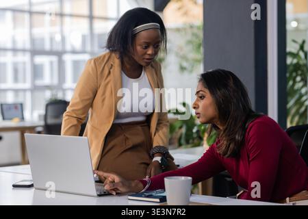Diverse female coworkers collaborating on laptop at modern office desk with notebook coffee mug Stock Photo