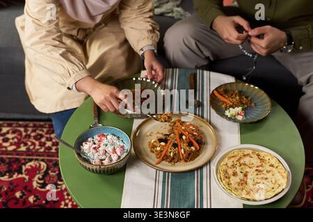 Muslim woman and Muslim man sitting at table sharing traditional meal, woman reaching for food while man holding prayer beads, dishes arranged on green table Stock Photo