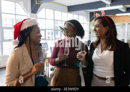 Diverse female co-workers clinking champagne flutes wearing suits, festive gear by desks in office Stock Photo