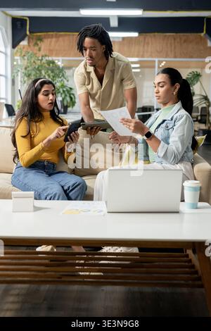Diverse coworkers collaborating around coffee table in office lounge using tablet and laptop Stock Photo