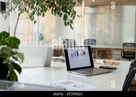 Silver laptop displaying colorful charts, resting next to smartphone on white desk in modern office Stock Photo