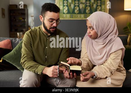 Young adult Muslim man holding prayer beads sitting with young adult woman wearing hijab, reading from small religious book together on sofa in living room Stock Photo