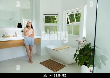 Leaning woman wearing tank top and light blue shorts at wood vanity, tub, plant, copy space Stock Photo