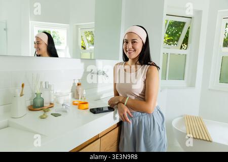 Asian woman standing and leaning against vanity wearing headband in bathroom, checking smartphone Stock Photo