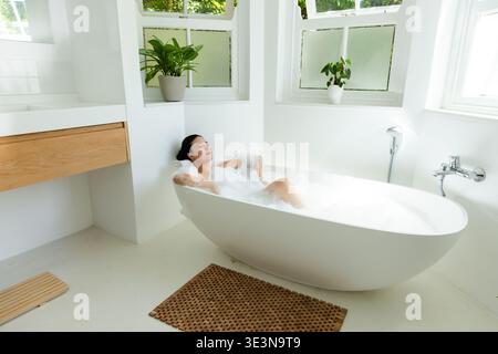 Woman reclining in freestanding tub filled with thick foam, closing eyes near frosted windows Stock Photo