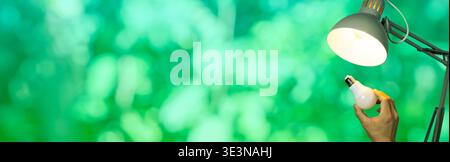 Hand holding an LED bulb near a lit desk lamp on a green bokeh background with copy space, representing energy saving, bulb replacement, electricity, Stock Photo