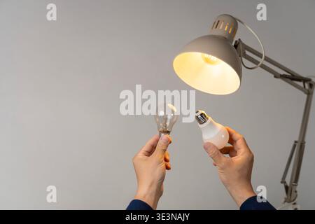 Hands holding incandescent and LED bulbs under a lit desk lamp on gray background, showing energy saving, lighting upgrade, replacement, comparison, a Stock Photo