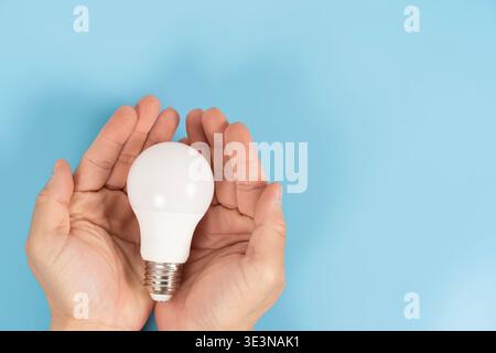 Close-up of hands holding an LED light bulb on blue background with copy space, symbolizing energy saving, protection, sustainable electricity, home l Stock Photo