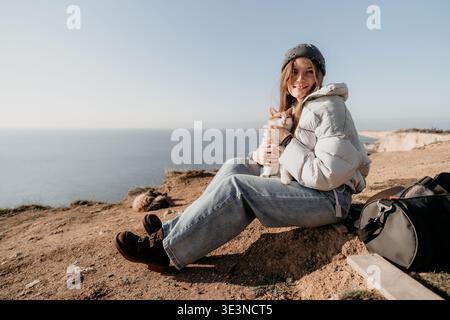 Woman cat travel sits on a scenic cliff edge holding her ginger pet feline companion while enjoying coastal view Stock Photo