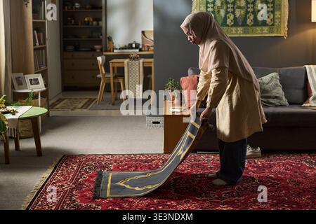Young Muslim woman preparing prayer rug in living room, standing on patterned carpet, arranging mat with both hands, traditional decor visible in background Stock Photo