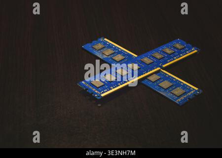 A high-angle close-up shot of two blue computer RAM memory sticks crossed to form an X shape. The modules are resting on a dark wood grain background, Stock Photo