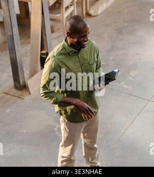 African American mature man in green shirt, beige trousers holding tablet and gesturing in woodshop. Woodworking, workshop, carpentry, industrial, con Stock Photo