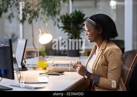 African American woman smiling while working on laptop at office desk with plant, copy space Stock Photo