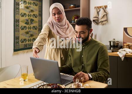 Young adult Muslim woman standing beside young adult Muslim man sitting at table using laptop, woman gesturing toward screen while man typing on keyboard Stock Photo