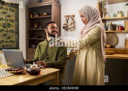 Young adult Muslim man sitting at desk using laptop, smiling at young adult Muslim woman standing beside him gesturing with hand in modern kitchen setting Stock Photo
