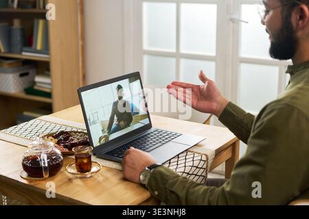 Young Muslim man with beard sitting at table gesturing while having video call with young adult Muslim man on laptop, tea and dates on table, bookshelf in background Stock Photo