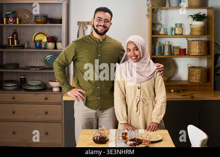 Young adult Middle Eastern man standing next to young adult Middle Eastern woman wearing hijab, both smiling and posing together in kitchen, traditional food and tea on table in front Stock Photo