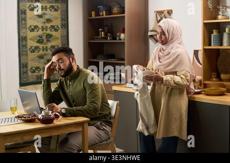 Young Muslim man sitting at table using laptop looking stressed, Muslim woman standing nearby holding plate and towel talking to him in modern kitchen Stock Photo