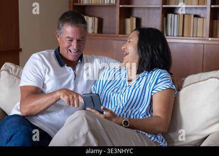 Diverse couple laughing and pointing at smartphone while sitting on sofa in living room Stock Photo