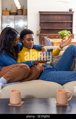 African American and Indian women sitting at home on sofa wearing yellow turtleneck, sharing phone Stock Photo