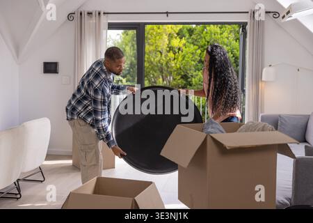 Couple carrying large round black tray while unpacking cardboard boxes in living room with linens Stock Photo