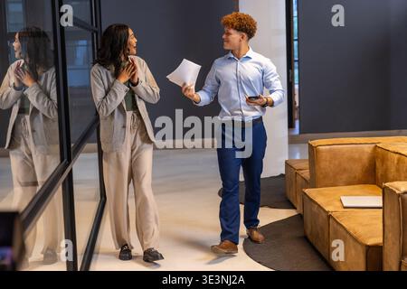 Diverse coworkers walking in office hallway while discussing papers and smartphone by glass wall Stock Photo