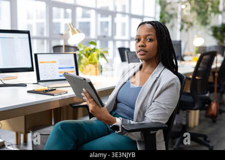 African American woman holding tablet and analyzing charts on monitors at open-plan office desk Stock Photo