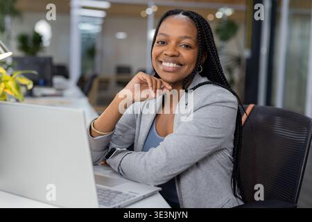African American woman wearing grey blazer while smiling at modern office desk with silver laptop Stock Photo