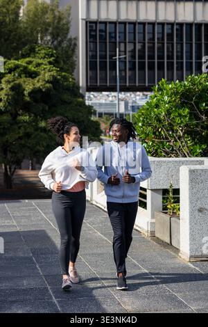 Diverse couple jogging on urban walkway wearing fitness tracker, passing concrete planters Stock Photo