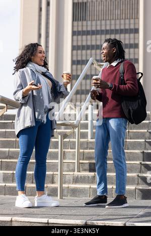 Diverse friends standing on steps at office entrance, gesturing with coffee cups, smartphone Stock Photo