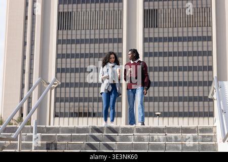 Diverse students walking down campus stairs holding coffee cups, backpacks with smartphone Stock Photo