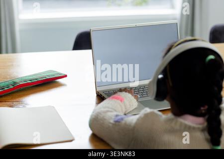 Child typing on laptop at home dining table near window, with notebook and patterned pencil case Stock Photo