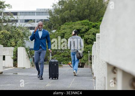 African american man in suit walking on office path pulling suitcase talking on phone Stock Photo