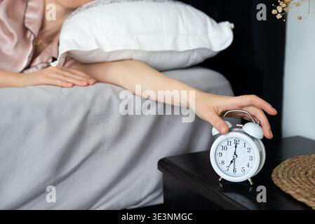 A woman in a pink satin top lies in bed, reaching for a white analog alarm clock on a black bedside table with a woven mat, morning light peeking through. Stock Photo