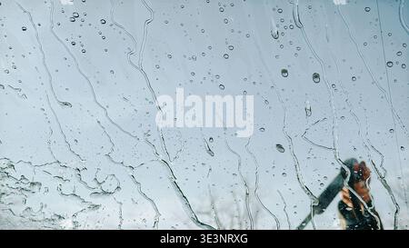 Close-up of water streaks and droplets on glass with a blurred hand using a pressure washer to clean a car. Dynamic scene ideal for car wash, maintena Stock Photo