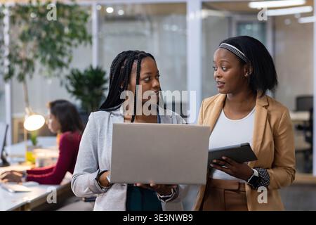 Two African American women colleagues reviewing data and typing on laptop and tablet in office Stock Photo