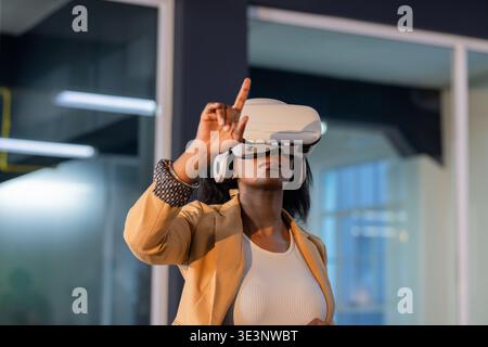 Woman wearing white VR headset and wristband, pointing finger, selecting virtual object in office Stock Photo