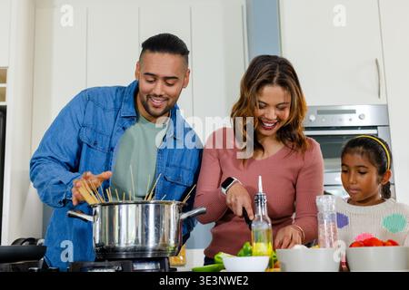 Family cooking in bright home kitchen stirring pot slicing vegetables and adding oil Stock Photo