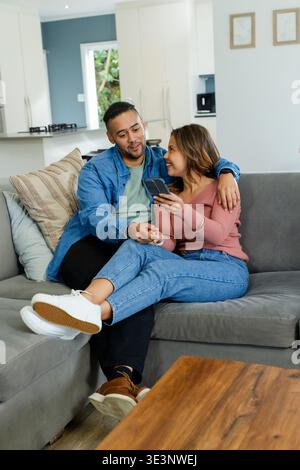 Couple sitting on grey sectional sofa with throw pillows in open living area, looking at smartphone Stock Photo