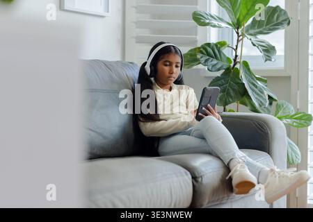 Asian school-age girl sitting on gray leather couch in living room holding smartphone, copy space Stock Photo