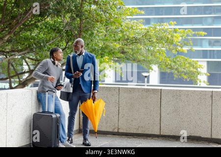 Diverse coworkers standing on terrace under tree holding tablet and suitcase Stock Photo