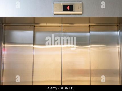 Closed stainless steel elevator door with digital floor display showing number four in red LED lights, representing modern building infrastructure and Stock Photo