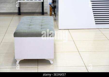 Light gray tufted upholstered bench with white wooden base placed on tiled floor against brick wall, showcasing modern functional seating design Stock Photo