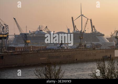 Deutschland, Hamburg: Kreuzfahrtschiff AIDAnova im Trockendock von BlohmVoss, massive Werftanlage im Vordergrund mit Dockmauern, Kränen und technischer Infrastruktur, im Hintergrund Hafenkräne und Windkraftanlage als Symbol für Industrie und Energiewandel, Teil der Flotte von AIDA Cruises, LNG-betriebenes Kreuzfahrtschiff mit charakteristischem AIDA-Kussmund am Bug, Hamburger Hafen als industrielles Zentrum und einer der größten Seehäfen Europas, Kontrast zwischen moderner Kreuzfahrtindustrie und klassischer Schiffswerft, warmes Abendlicht mit leichtem Dunst, urbane Hafenlandschaft, Logistik, Stock Photo