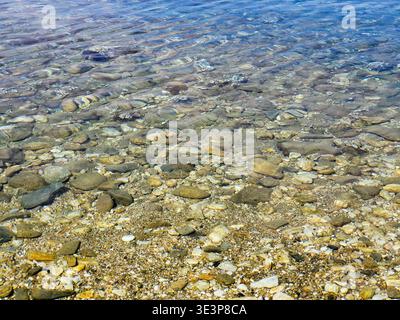 Crystal-clear shallow water gently rippling over a pebble beach, sunlit stones visible beneath the surface creating textured patterns and a calm, peac Stock Photo