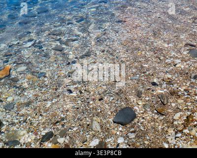 Crystal-clear shallow water gently rippling over a pebble beach, sunlit stones visible beneath the surface creating textured patterns and a calm, peac Stock Photo
