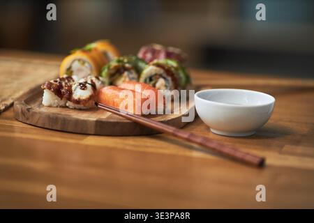 Assorted sushi rolls and nigiri arranged on wooden board with wooden chopsticks resting beside small bowl, showcasing traditional Asian cuisine and cultural dining elements Stock Photo