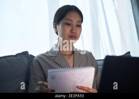 Asian young adult woman sitting on sofa holding notepad and looking at laptop screen, engaging in remote work or online learning, focused expression, cultural context visible Stock Photo
