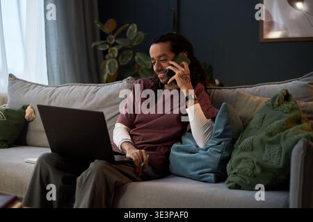 Young adult Asian man sitting on sofa multitasking with laptop and smartphone, smiling while talking on phone and working remotely in modern living room with decorative pillows Stock Photo